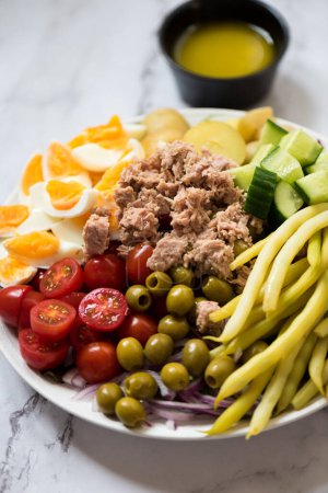 Photo for Nicoise Salad - French style salad with eggs, tuna, cherry tomatoes, green olives, potatoes, green beans and cucumber on big plate on white marble background. Tasty food recipe cook book. - Royalty Free Image