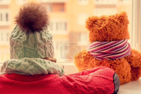 Téléchargez les photos : La fille dans une casquette à une fenêtre avec un ours en peluche. La femme dans un chapeau chaud et un ourson jouet regardent par la fenêtre. J'attends le printemps. Le concept d'attendre l'arrivée du printemps. gros plan - en image libre de droit