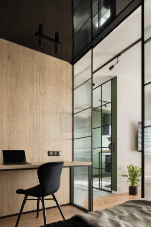 Foto de Moderna oficina en casa con silla negra simple y escritorio de madera en la habitación con elegante pared de vidrio reforzado - Imagen libre de derechos