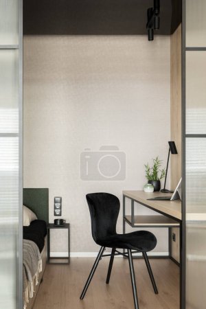 Foto de Elegante silla negra en la sala de oficina en casa con escritorio de madera, cama y puerta de vidrio reforzado - Imagen libre de derechos