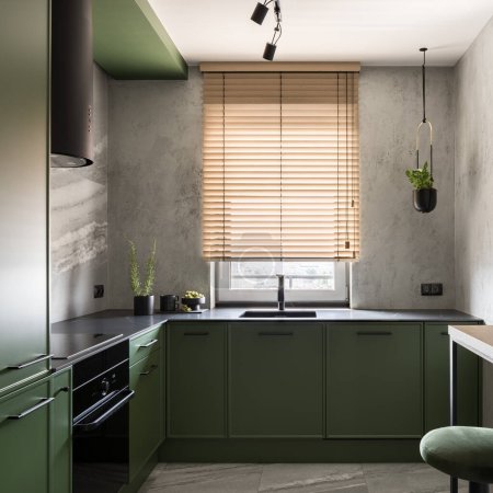 Spacious and modern kitchen with green furniture, black oven, kitchen hood and countertops, concrete on walls and window with wooden blinds