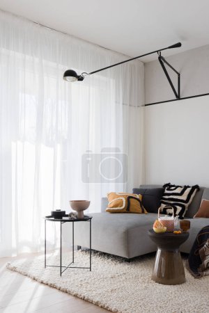 Photo for Eclectic and bright living room with big window behind white curtain, elegant sofa with decorative pillows, coffee tables with decorations and modern black lamp - Royalty Free Image