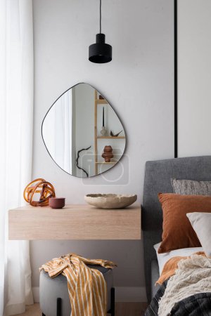 Photo for Modern, irregular shaped mirror above simple, wooden dressing table next to cozy bed in bedroom - Royalty Free Image