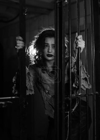 Photo for Brunette girl in Halloween style. A woman in prison, a scary scene. Terrible darkness, torn clothes on the girl. Hostage in a cage, depression, suffering, crime. Black Carnival, in the basement. - Royalty Free Image