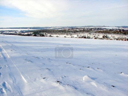 Blick von der Spitze eines schneebedeckten Hügels auf die Privathäuser der Siedlung unter einem klaren blau-weißen Himmel am Horizont.