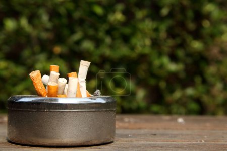 Beside view of round shape stainless ashtray is on wooden table and a cigarette in it and blur tree background.