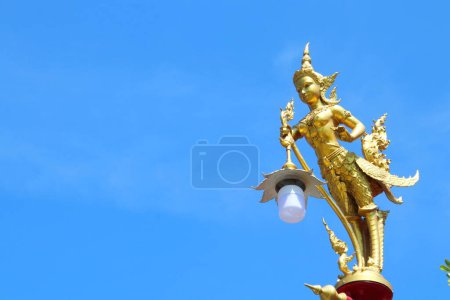 Photo for Golden color painted Kinnaree statue holding lamp at the top of pillar and bright blue sky background. Kinnaree is female bird with human head in ancient Thai tale. - Royalty Free Image
