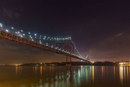 Foto de Puente rojo iluminado 25 de Abril Puente que cruza el río Tajo con la estatua de Cristo Rei durante la noche, Lisboa, Portugal - Imagen libre de derechos