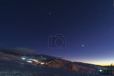 Snow covered mountains of Sierra Nevada near Granada with ski slope and view at IRAM radio telescope at twilight with stars on sky, Andalusia, Spain