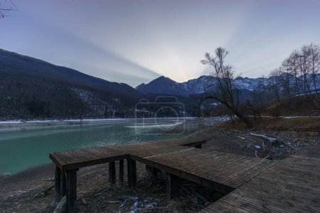 Winter sunset behind a mountain peak of a winter alpine landscape with wooden pier at reservoir lake at Barcis, Friuli-Venezia Giulia, Italy