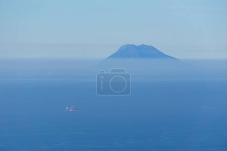 Téléchargez les photos : Volcan Stromboli lointain avec mer bleue et ciel par une journée brumeuse et ensoleillée à partir de Palmi, Calabre, Italie - en image libre de droit