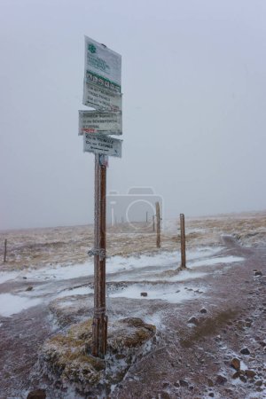 Ice covered trail marker post on Hohneck peak on GR5 hiking path at the Vosges Mountains during stormy weather conditions in winter, Alsace, France