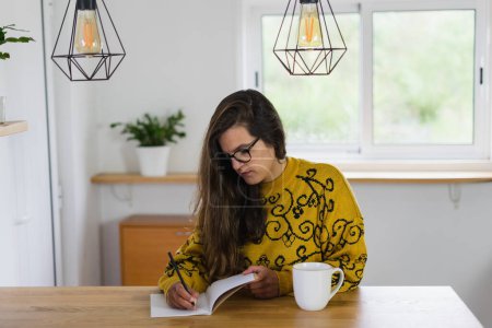 Foto de Mujer con gafas escribiendo en su cuaderno con una taza blanca a su lado en una encimera de madera. Foto de alta calidad - Imagen libre de derechos