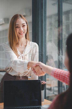Téléchargez les photos : Deux femmes d'affaires souriantes serrant la main en accord lors d'une réunion de bureau réussie, avec un ordinateur portable sur la table - en image libre de droit