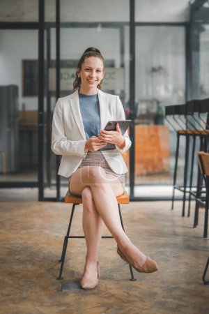Business analytics concept, Elegant businesswoman in a modern office setting sits on a high stool, holding a digital tablet with a pleasant smile