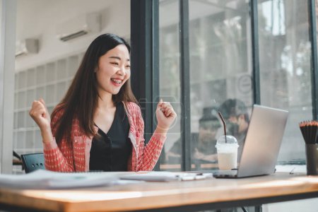 Business Analytics-Konzept, Eine begeisterte junge Frau feiert einen erfolgreichen Moment bei der Arbeit vor ihrem Laptop in einem lebendigen Bürobereich