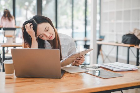Freiberufliches Konzept, besorgte asiatische Geschäftsfrau, die mit einer Hand auf dem Kopf Dokumente liest und in einem geschäftigen Café an einem Laptop arbeitet