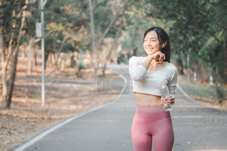 Photo for A thoughtful young athlete stands holding a water bottle on a park path, pausing for a moment of rest during her workout, surrounded by the calm of nature. - Royalty Free Image