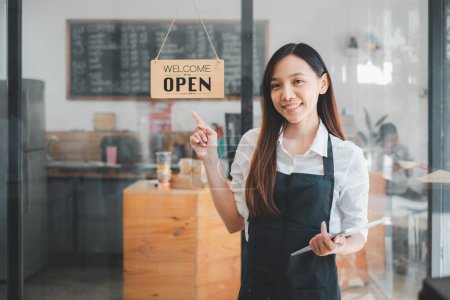 A cheerful cafe owner stands by the 'Welcome Open' sign at her storefront, tablet in hand, ready to greet customers into the warm atmosphere of her coffee shop.