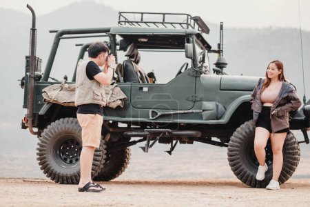 Man is focused on photographing a smiling woman who is leaning against a rugged off-road car.