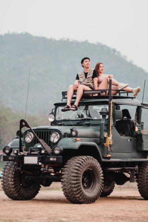 Adventurous couple is perched atop a rugged car, the woman pointing towards the horizon, ready for their next off-road exploration.