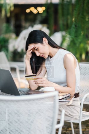 Mujer joven preocupada experimentando problemas con un pago en línea, sosteniendo una tarjeta de crédito y mirando la pantalla de un ordenador portátil en un café al aire libre.