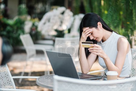 Mujer joven preocupada experimentando problemas con un pago en línea, sosteniendo una tarjeta de crédito y mirando la pantalla de un ordenador portátil en un café al aire libre.