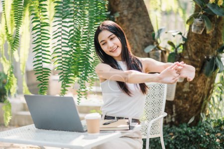 Relajada freelancer femenina estirando los brazos mientras toma un descanso del trabajo en su computadora portátil en un exuberante jardín cafetería.