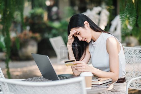 Worried young woman experiencing problems with an online payment, holding a credit card and looking at laptop screen at an outdoor cafe.