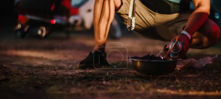 Camper se agacha para encender una parrilla de carbón al atardecer, con un brillo rojo que ilumina la atmósfera del camping.