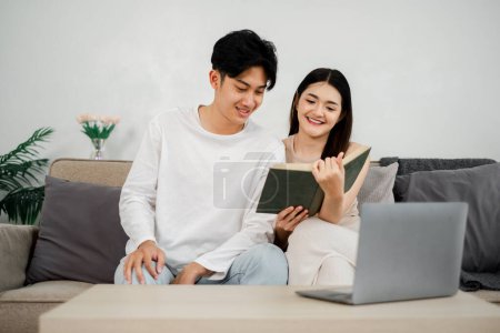 Photo for Couple sits comfortably on a couch, sharing a book, with a sense of closeness and a laptop waiting to be used. - Royalty Free Image