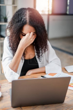 Mujer profesional con exceso de trabajo que muestra signos de estrés y dolor de cabeza mientras trabaja en su computadora portátil en una oficina moderna.