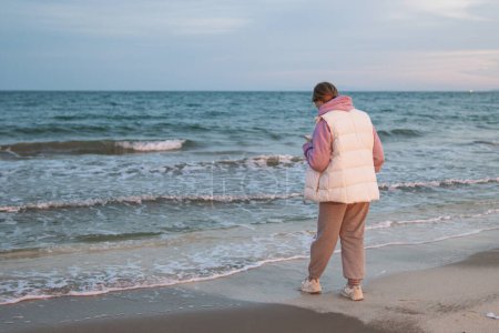 Téléchargez les photos : Une fille de marque européenne en vêtements décontractés se tient près de l'océan avec un téléphone dans ses mains est triste et regarde dans le téléphone. Photo de haute qualité - en image libre de droit