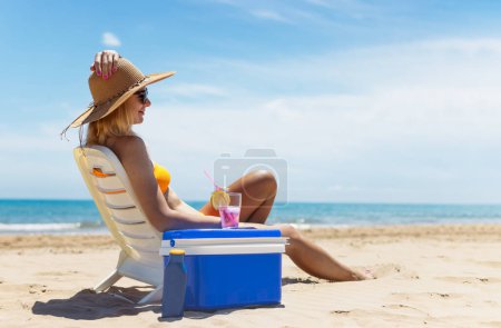 Téléchargez les photos : Heureuse jeune fille européenne boit un cocktail et se repose dans une chaise longue sur la plage, espace pour une inscription portable réfrigérateur bleu au bord de la mer. Photo de haute qualité - en image libre de droit