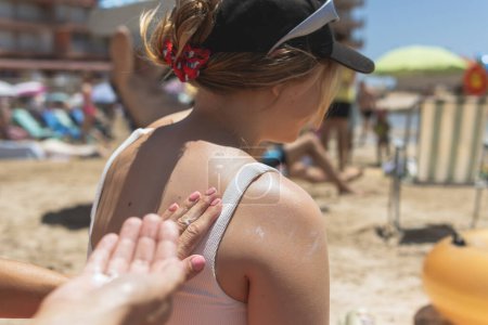 Vor dem Hintergrund des Strandes trägt eine Mutter liebevoll Sonnencreme auf den Rücken ihrer Tochter im Teenageralter auf und betont die Bedeutung des Sonnenschutzes während der sommerlichen Entspannung mit Kindern.