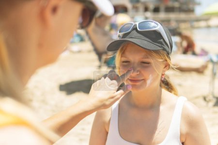 At the sunny beach, a mother lovingly applies sunscreen to her daughter's nose, exemplifying the essence of summer relaxation with children by the sea.