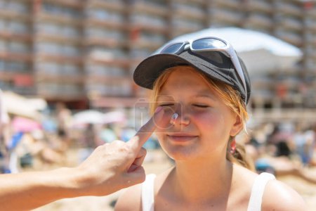 Against the backdrop of the summer beach, a mother tenderly applies sunscreen to her daughter's face as they sit in their swimsuits, embodying the essence of seaside relaxation and parental care.