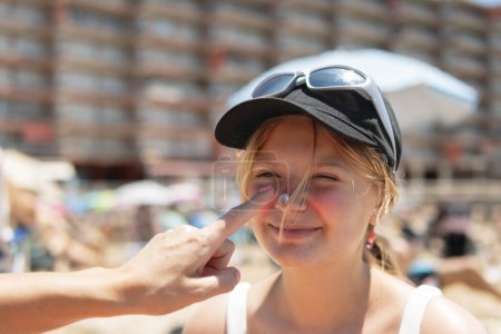 Against the backdrop of a sunny beach, a mother gently applies sunscreen to her daughter's face, both clad in swimsuits, embodying the essence of summer relaxation and sun protection.
