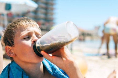 A boy enjoys a refreshing drink by the sea, his portrait framed against the beach backdrop, embodying the essence of summer relaxation and seaside enjoyment