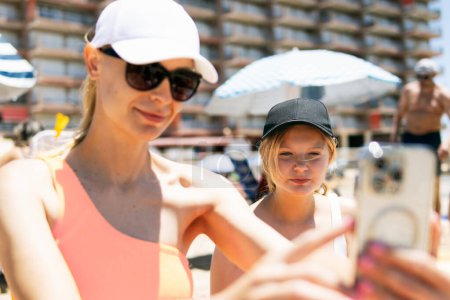 Eine Mutter und eine Tochter im Teenageralter, beide in Badeanzügen, machen vor der Kulisse eines sonnigen Strandes ein Selfie, das für sommerliche Bindung und Spaß steht.
