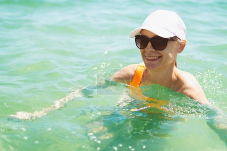 A happy woman in a swimsuit, sunglasses, and white cap swims in the sea during summer, epitomizing the joy of oceanic leisure.