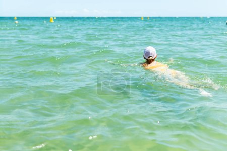 A happy woman in a swimsuit, and white cap swims in the sea during summer, capturing the essence of carefree ocean enjoyment.