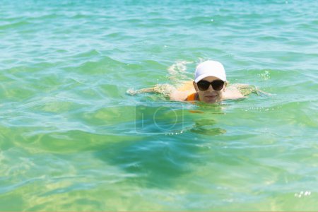 A happy woman in a swimsuit, sunglasses, and white cap enjoys a swim in the sea during summer, capturing the essence of carefree oceanic bliss.