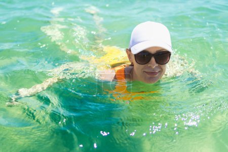 A happy woman in a swimsuit, sunglasses, and white cap enjoys a swim in the sea during summer, capturing the essence of carefree oceanic bliss.