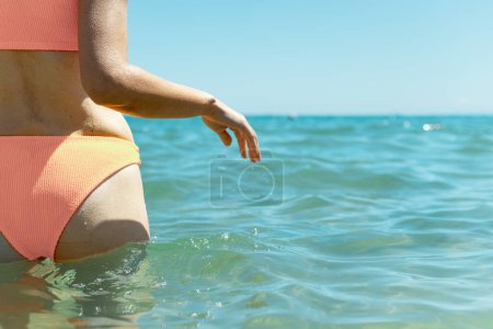 Una chica delgada, mirando lejos de la cámara, se para en el mar azul vistiendo un traje de baño, personificando la serena belleza junto a la playa.