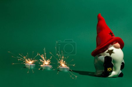 Téléchargez les photos : Figurine de Noël en céramique de gnome avec chapeau rouge et barbe blanche et bougies d'arbre avec scintillement sur fond vert. - en image libre de droit