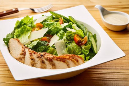 Herb Chicken Caesar Salat mit Kirschtomaten, Gurken, Salatblättern und Hähnchenbrustscheibe serviert in Schale isoliert auf Holztisch mit Rohkost Draufsicht auf super gesunde Vorspeisengerichte