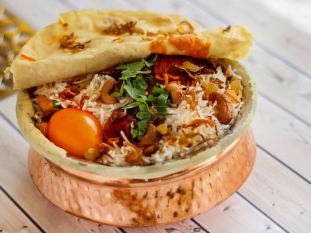 Indian Chicken Biryani with tomato, coriander, cashew nut and bread dum served in copper pot dish isolated on wooden table top view arabic spicy food