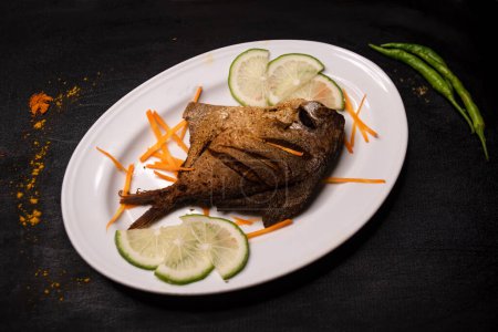 Gebratener Pomfret Fish oder Rupchanda Fry mit Limettenscheibe serviert in Schale isoliert auf dunklem Hintergrund Draufsicht indische Gewürze, Bangladeshi und pakistanisches Essen
