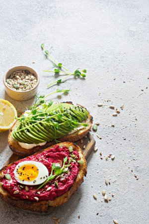 Téléchargez les photos : Whole grain toast with cream cheese, avocado, beetroot hummus, seeds, egg and microgreens (sprouted pea sprouts) on a light gray background. Healthy food concept. - en image libre de droit